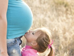 A young girl lovingly kisses her pregnant mother's belly outdoors, symbolizing family affection.
